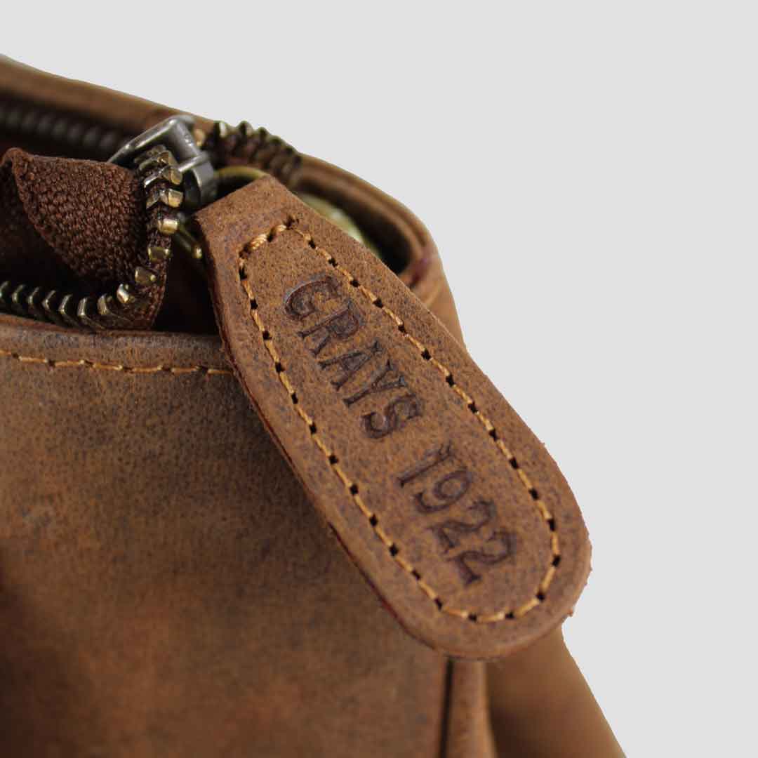 Close-up of a brown leather bag with 'Grays 1922' embossed logo on the zip pull